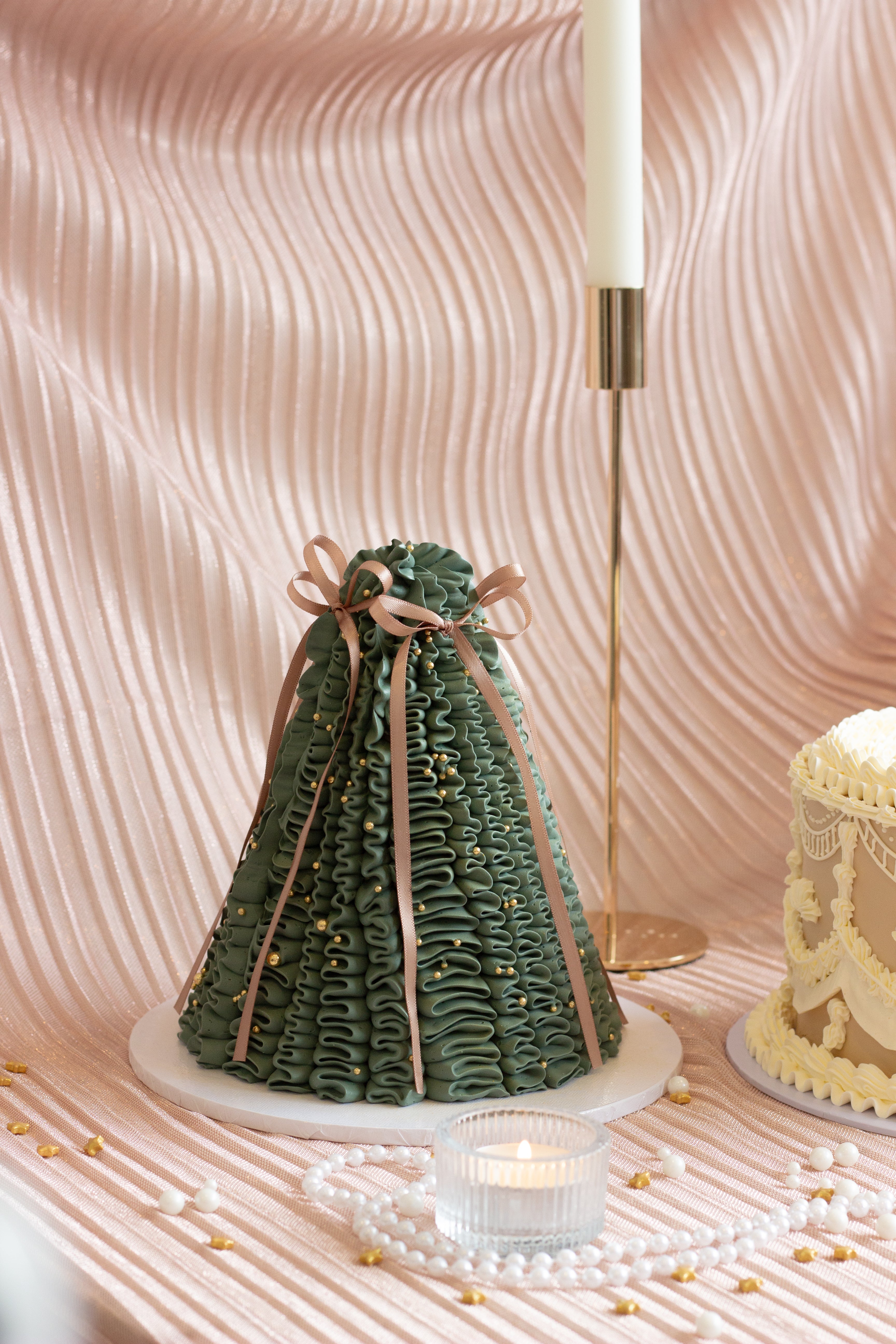 A set of four Christmas cakes displayed on soft pink draped fabric, including green tree cakes, a vintage ivory cake, and a pink cherry-topped tree cake with candles and pearls.

Tall dark green pleated Christmas tree cake decorated with gold pearls and a red bow. Cake Melbourne. Christmas cake Melbourne.
