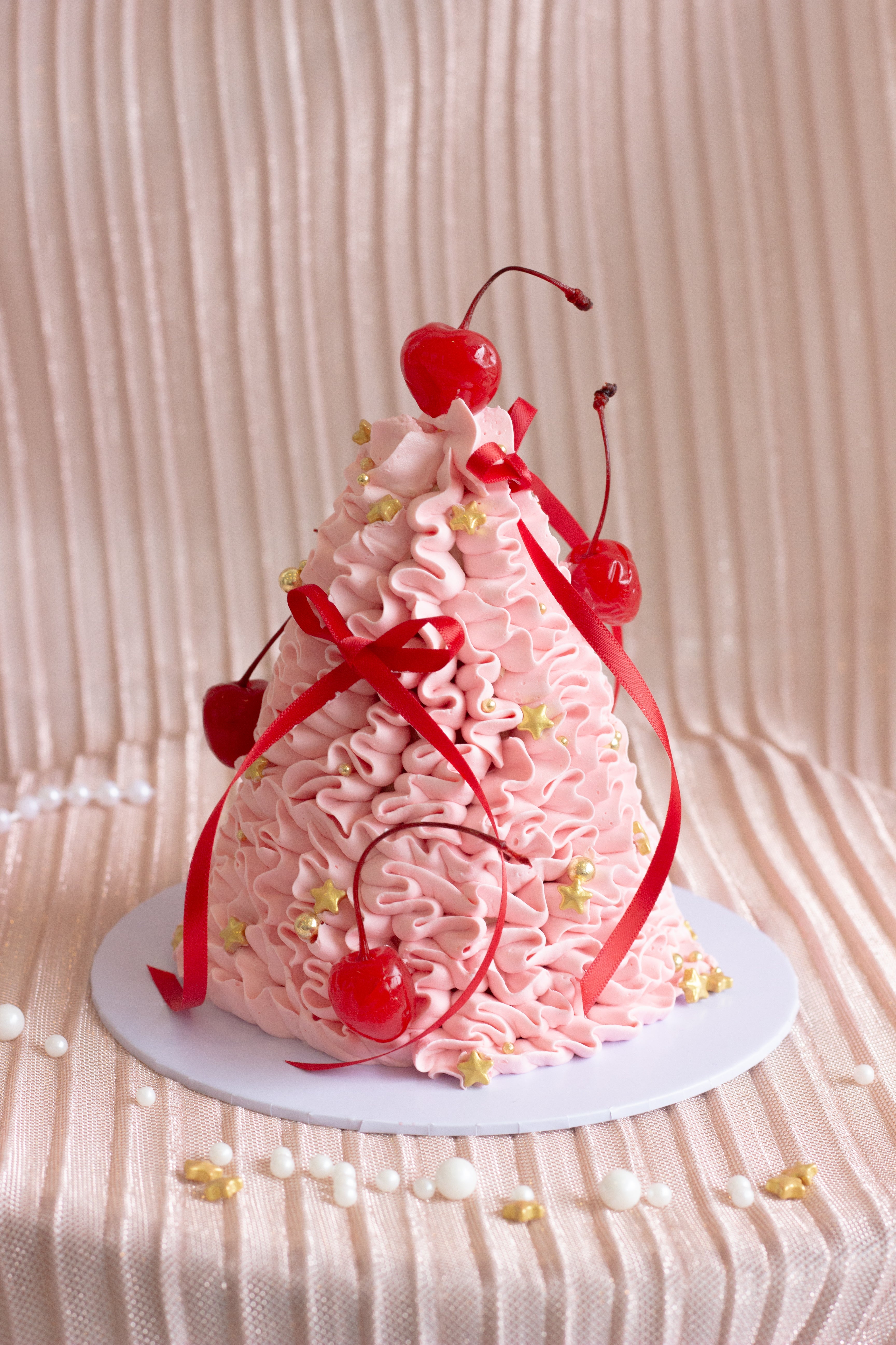 Pink Christmas tree shaped cake with expressive buttercream piping, red ribbons, and cherries on top.