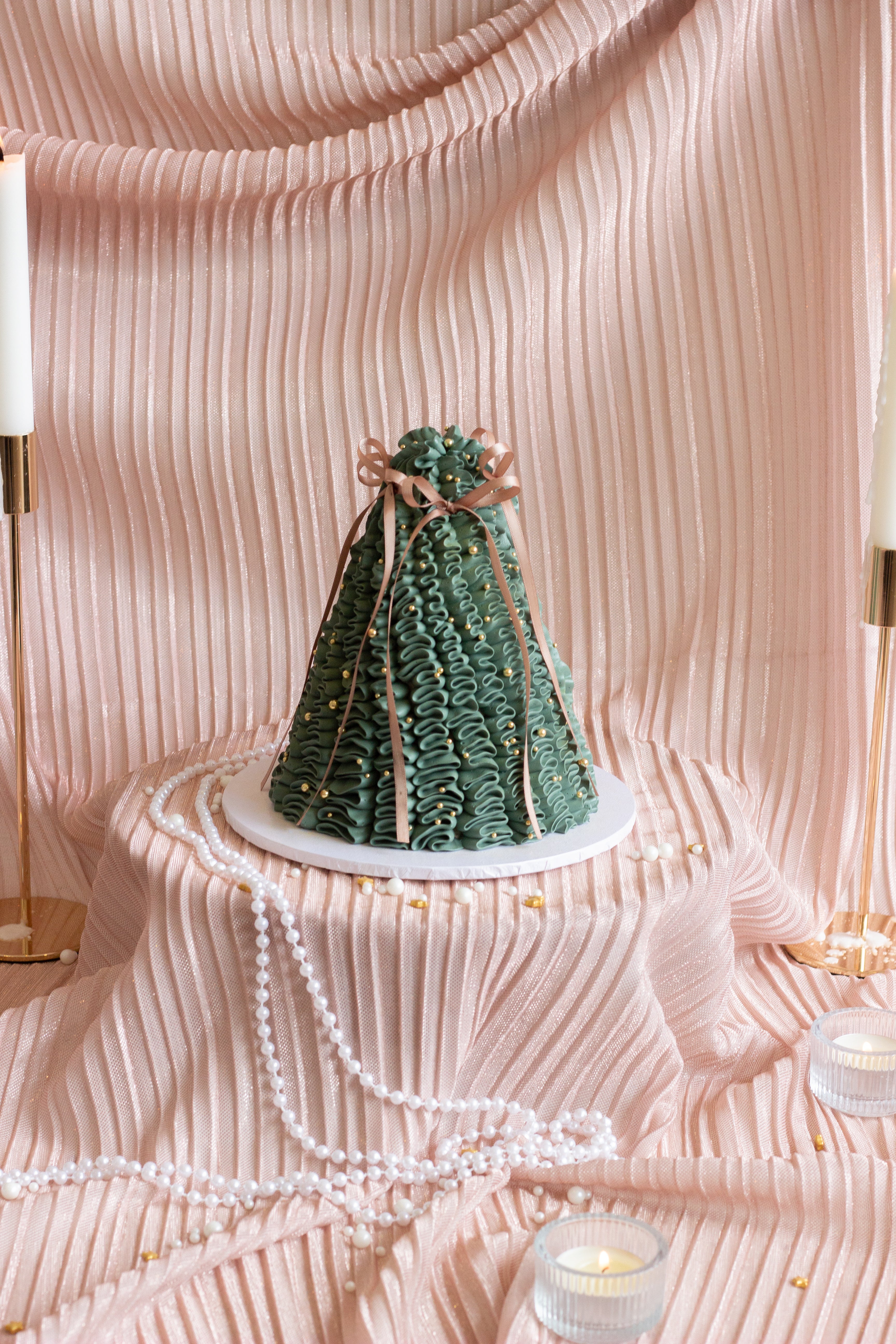 A set of four Christmas cakes displayed on soft pink draped fabric, including green tree cakes, a vintage ivory cake, and a pink cherry-topped tree cake with candles and pearls.

Tall dark green pleated Christmas tree cake decorated with gold pearls and a red bow. Cake Melbourne. Christmas cake Melbourne.