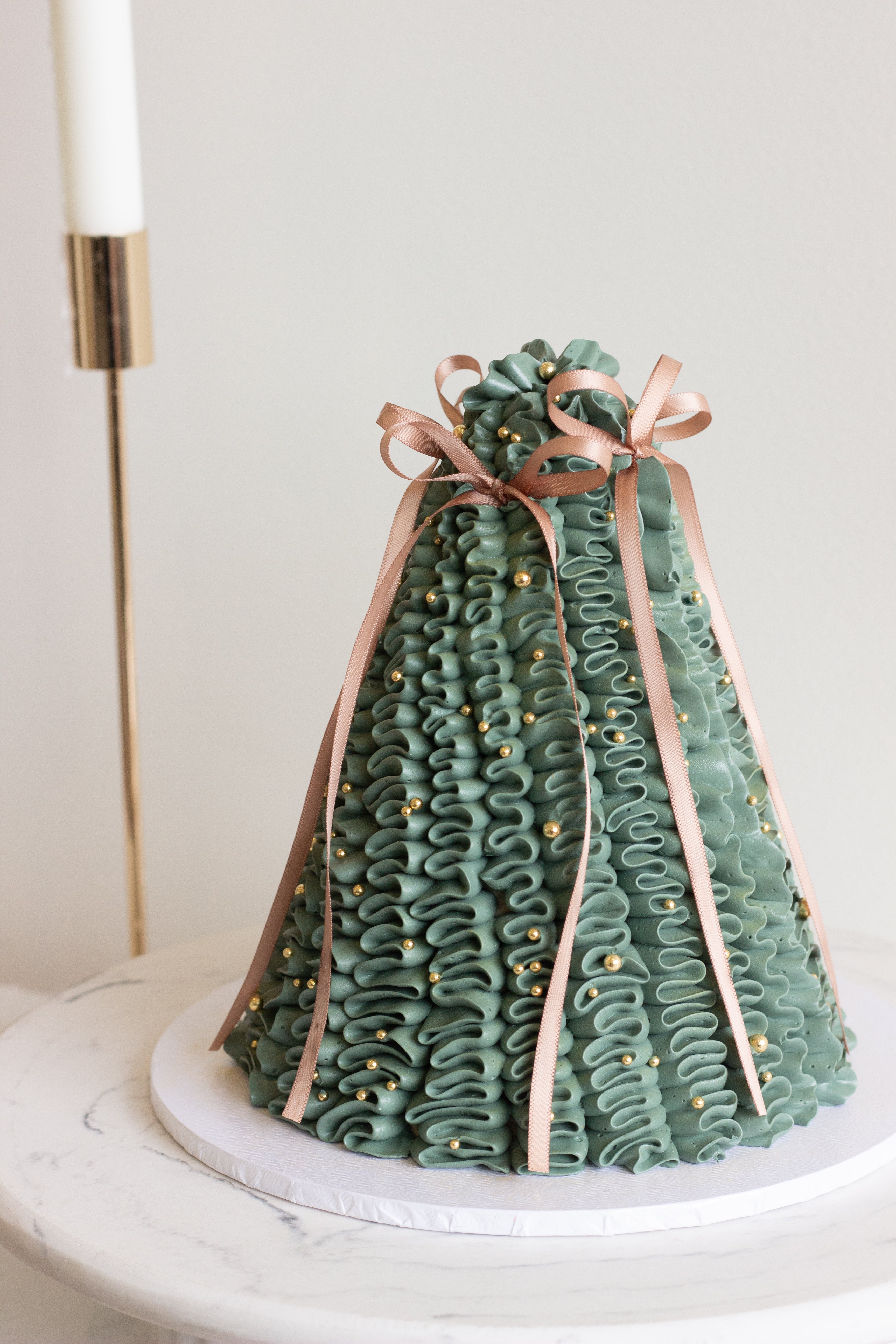 A set of four Christmas cakes displayed on soft pink draped fabric, including green tree cakes, a vintage ivory cake, and a pink cherry-topped tree cake with candles and pearls.

Tall dark green pleated Christmas tree cake decorated with gold pearls and a red bow. Cake Melbourne. Christmas cake Melbourne.